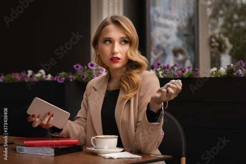 Charming young woman with long blonde hair holding the phone in her hands while sitting in a street cafe