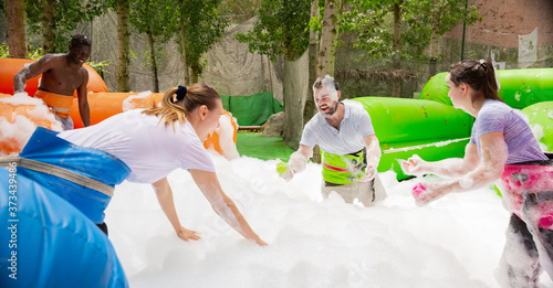 Cheerful friends having fun in outdoor amusement park, picking up balls in inflatable pool full of foam © JackF