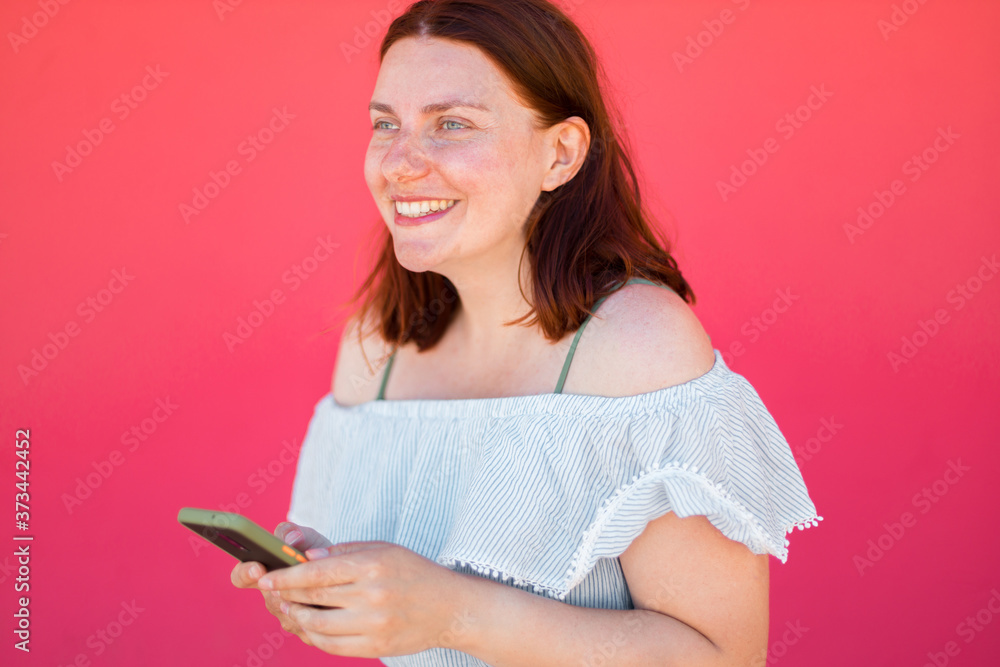 Caucasian beautiful young girl smiling and using smartphone on pink wall background with copy space Caucasian beautiful young girl smiling and using smartphone on pink wall background with copy space