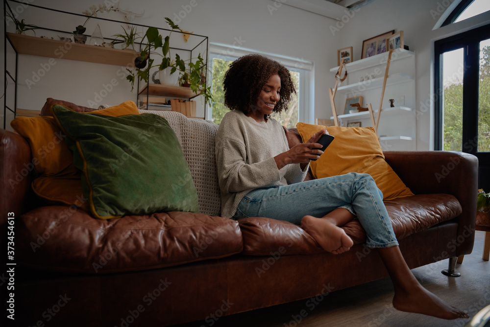 Obraz premium Smiling african woman sitting on comfortable couch using smartphone at home during lockdown