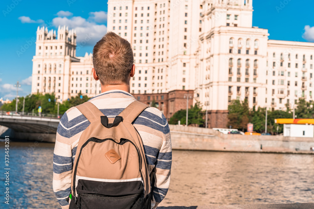 A young man with a backpack walks along the embankment with a view of the Stalin high-rise and the Moscow river, sights of Moscow