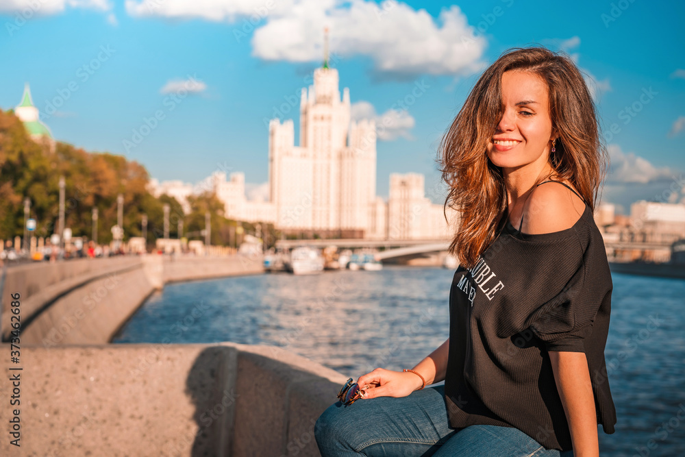 Moscow / Russia - 15 Aug 2020: Portrait of a young brunette woman in a jacket with an inscription with the background of the famous high-rise building and the Moscow river on a Sunny summer day
