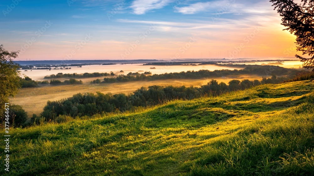 Beautiful morning landscape with green grass on the hill, blue sky and misty valley in the background