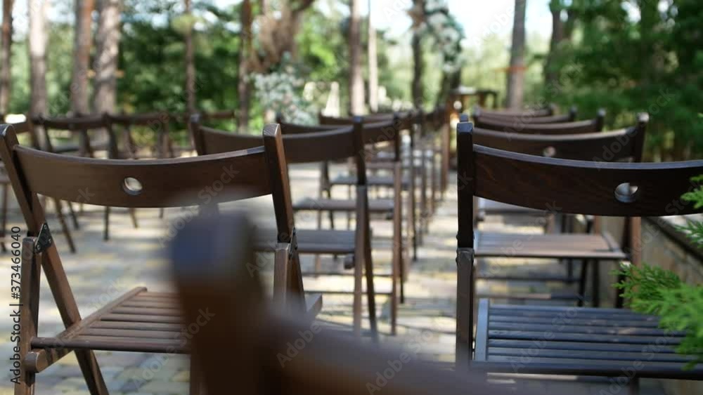 A large number of brown wooden chairs are lined up against the backdrop of the wedding decor. Empty seats for guests on the background of the wedding arch. A large number of brown wooden chairs are lined up against the backdrop of the wedding decor. Empty seats for guests on the background of the wedding arch.