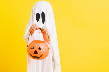 Funny Halloween Kid Concept, little cute child with white dressed costume halloween ghost scary he holding orange pumpkin ghost on hand, studio shot yellow on white background