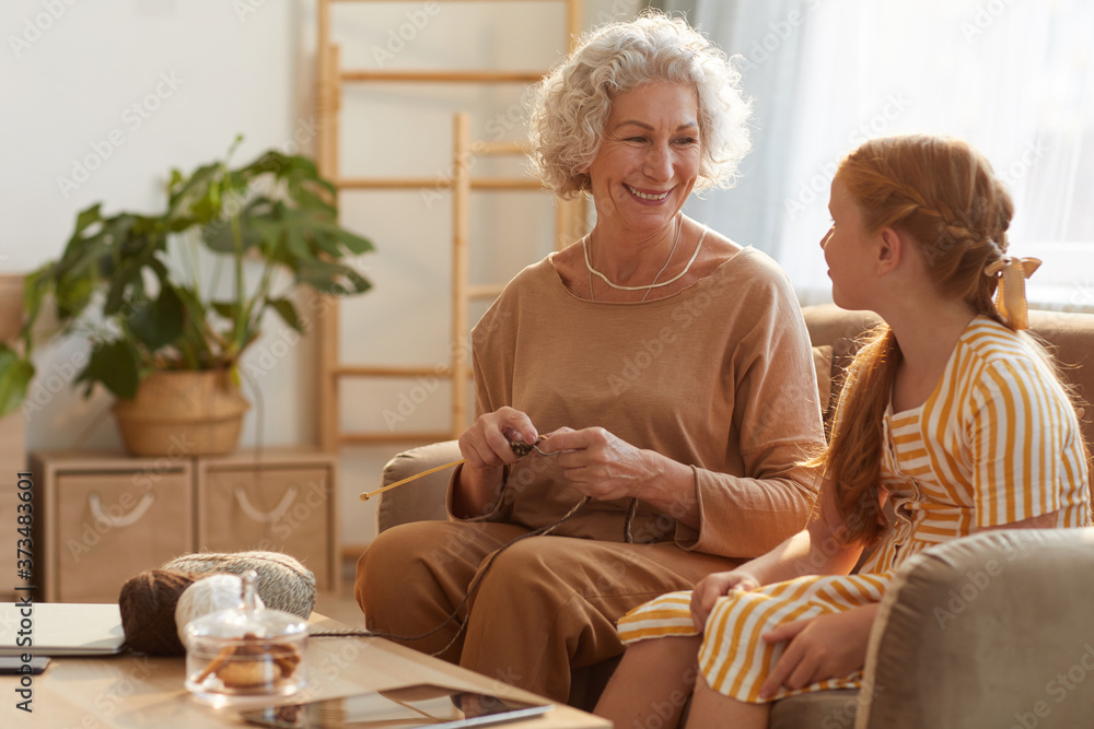 Warm toned portrait of smiling senior woman knitting with cute girl watching her in cozy home lit by sunlight, copy space Warm toned portrait of smiling senior woman knitting with cute girl watching her in cozy home lit by sunlight, copy space