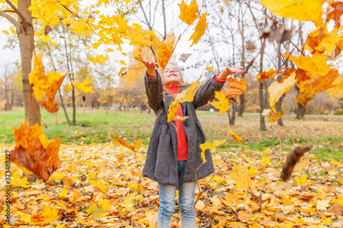 Happy young girl playing under falling yellow leaves in beautiful autumn park on nature walks outdoors. Little child throws up autumn orange maple leaves.