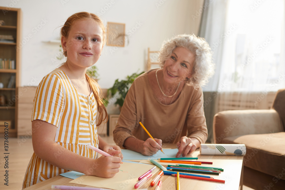 Warm toned portrait of cute red haired girl smiling cheerfully at camera while drawing together with grandma enjoying time at home, copy space Warm toned portrait of cute red haired girl smiling cheerfully at camera while drawing together with grandma enjoying time at home, copy space