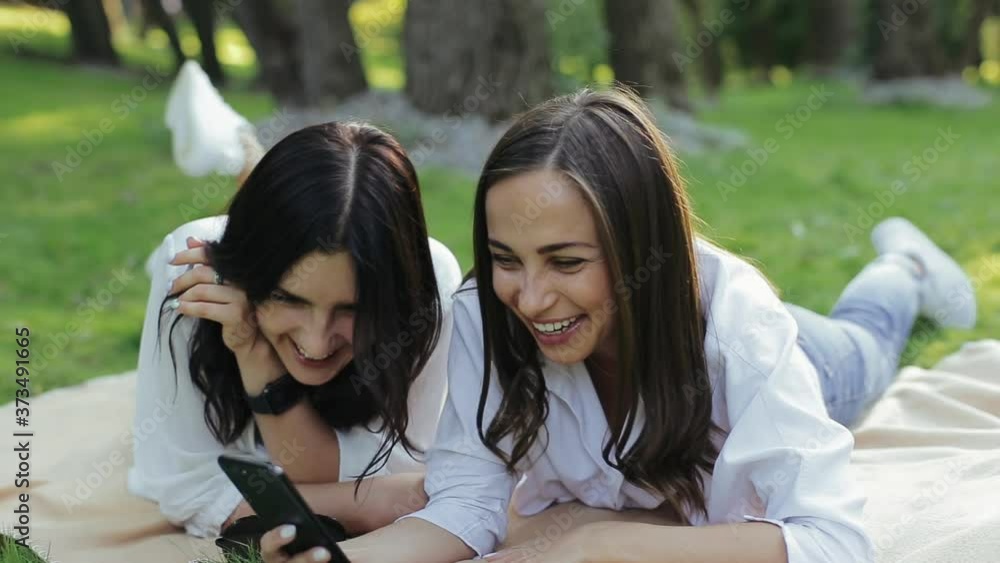 Two cute funny girlfriends looking in smartphone, sings song in karaoke, having fun lying on grass in cozy park among trees. Pair happy beautiful young brunettes watching new video clip and singing Two cute funny girlfriends looking in smartphone, sings song in karaoke, having fun lying on grass in cozy park among trees. Pair happy beautiful young brunettes watching new video clip and singing