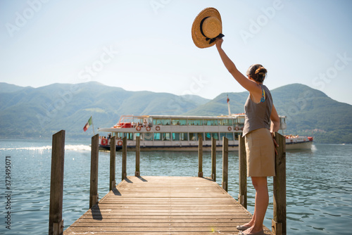 Woman Standing on a Pier and Greeting with Her Straw Hat a Passenger Ship on Alpine Lake Maggiore with Mountain in Ticino, Switzerland.