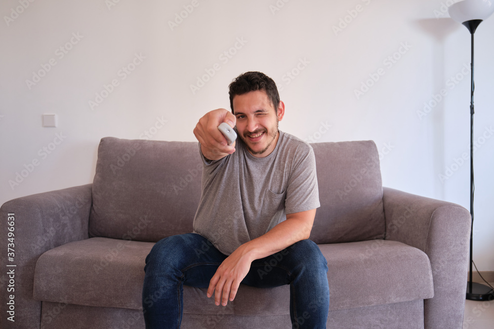 Portrait of a smiling young man sitting on a sofa and changing TV channels with the remote control on his hands.