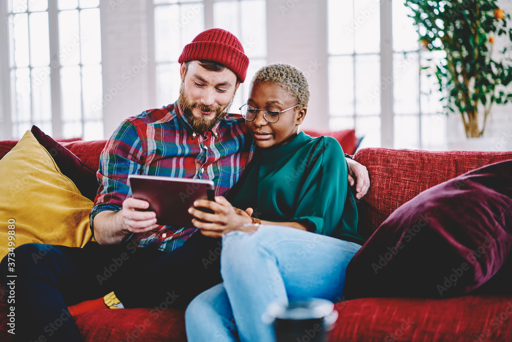 Young diverse marriage relaxing on sofa choosing furniture for own flat during internet shopping via digital tablet, millennial couple in love watching online movie via touchpad streams app