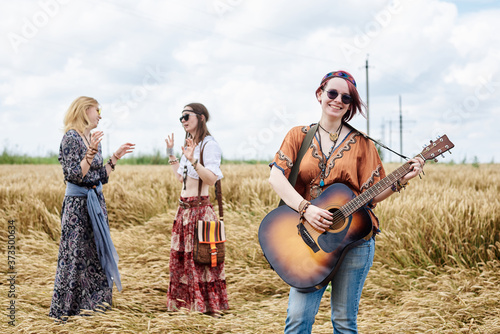 Three hippie women, wearing boho style clothes, dancing in the wheat field, playing guitar, laughing, Female friends, traveling together in countryside. Eco tourism concept. Summer leisure time