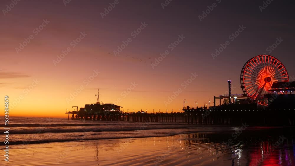 Twilight waves against classic illuminated ferris wheel, amusement park on pier in Santa Monica pacific ocean beach resort. Summertime iconic symbol of California glowing in dusk, Los Angeles, CA USA Twilight waves against classic illuminated ferris wheel, amusement park on pier in Santa Monica pacific ocean beach resort. Summertime iconic symbol of California glowing in dusk, Los Angeles, CA USA