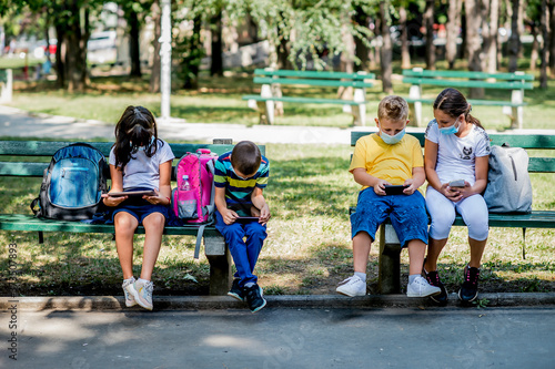 Back to school during quarantine. Children with backpacks wearing protective face mask and using mobile phones and digital tablets.