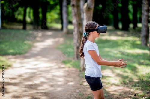 Fototapeta Naklejka Na Ścianę i Meble -  School girl in white t shirts having fun with virtual reality headset outdoor in the park