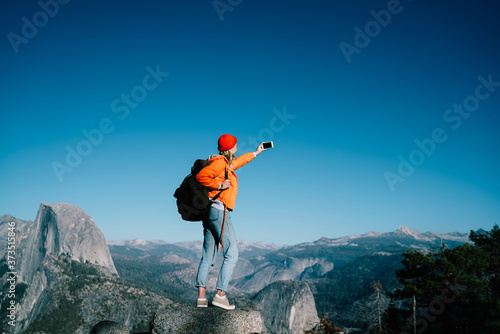 Young female traveler in orange jacket using mobile phone for taking photo on scenic natural view on active leisure, woman hipster wanderlust standing on mountain shooting video for blog
