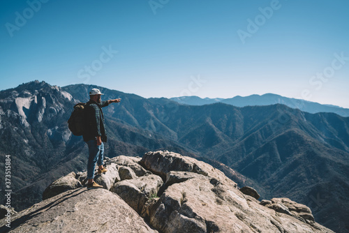 Caucasian male traveler with backpack standing on rock top pointing on horizon and high mountains on hiking tour, hipster guy explorer showing direction having active leisure time adventure
