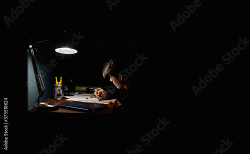 Teenage boy drawing at a desk in a dark room by lamp light.