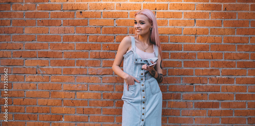 Carefree female millennial holding modern cellphone device standing at urbanity with promotional brick wall on background, happy hipster girl dressed in jeans sundress smiling at publicity area