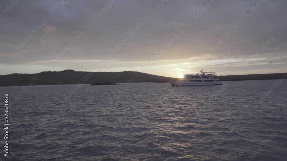 A cruise is anchored near an island of the Galapagos islands during sunset. The calm ocean surrounds the beach that has some clouds over it. A cruise is anchored near an island of the Galapagos islands during sunset. The calm ocean surrounds the beach that has some clouds over it.