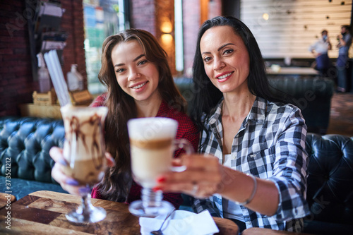 Casual happy smiling modern mother and daughter having coffee break together and enjoying a coffee cocktails in a coffee shop