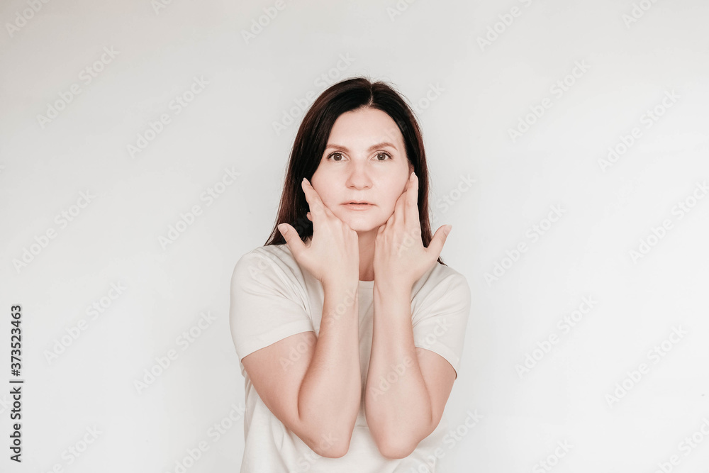 Beautiful brunette woman keeps hands on cheek, dressed in casual white t shirt, has minimal makeup, poses indoor on a white background. Copy, empty space for text Beautiful brunette woman keeps hands on cheek, dressed in casual white t shirt, has minimal makeup, poses indoor on a white background. Copy, empty space for text