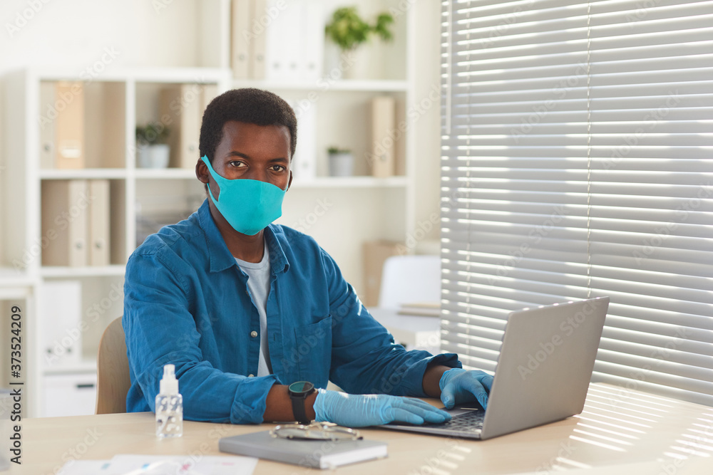 Portrait of young African-American man wearing mask and gloves looking at camera while working at desk in post pandemic office, copy space Portrait of young African-American man wearing mask and gloves looking at camera while working at desk in post pandemic office, copy space