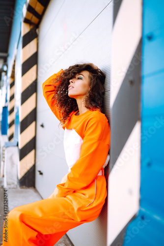 Portrait of stylish woman with curly hair in an orange suit. Young African American woman hip-hop dancer at sunset. Sport, dancing and urban culture concept. 
