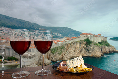 Two glasses of wine with charcuterie assortment against view of Dubrovnik, Croatia. Glass of red wine with different snacks - plate with ham, sliced, blue cheese. Romantic celebration.