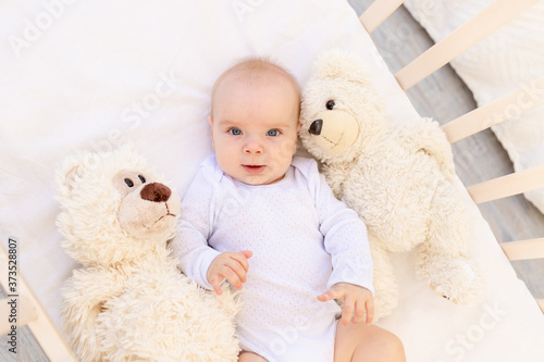 portrait of a small child a girl 6 months old in a white bodysuit lying on her back in a child's bed with soft toys bears