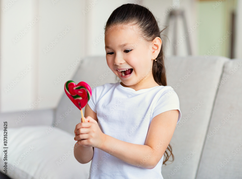 Excited Kid Girl Holding Lollipop Sitting On Sofa At Home Excited Kid Girl Holding Lollipop Sitting On Sofa At Home