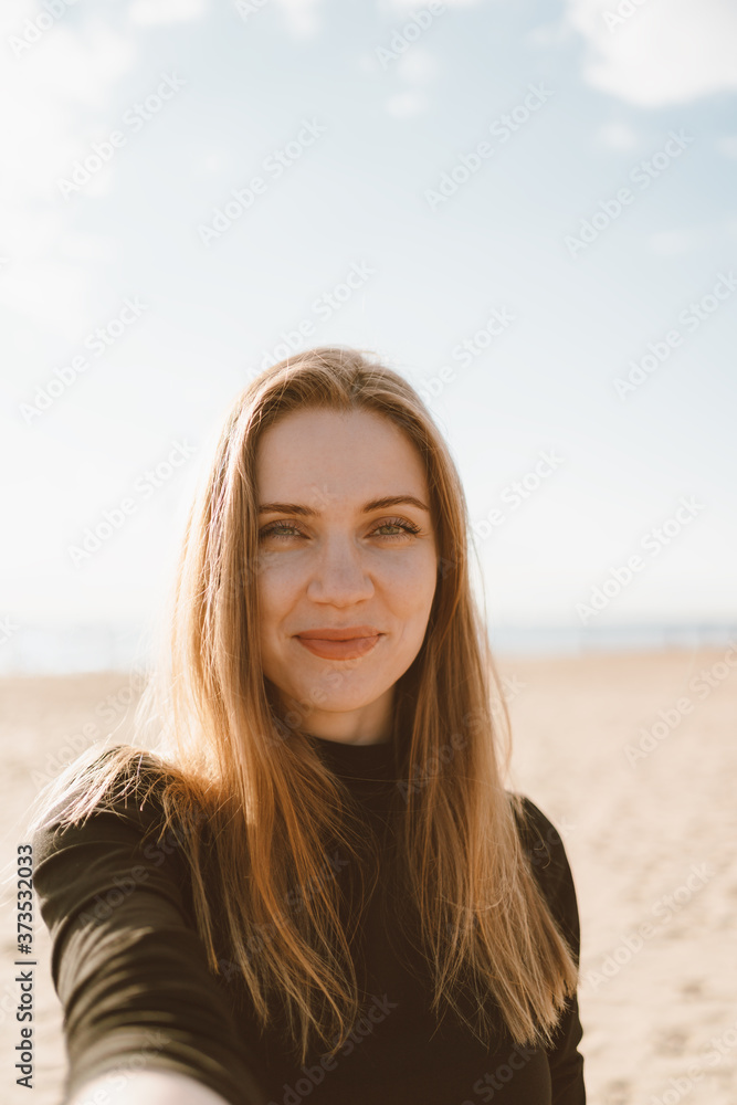 Pretty female with long hair, blonde takes selfie on mobile phone on sandy beach in summer evening. Beautiful woman looking at camera and smiling in sunlight in ocean or sea coastline Pretty female with long hair, blonde takes selfie on mobile phone on sandy beach in summer evening. Beautiful woman looking at camera and smiling in sunlight in ocean or sea coastline
