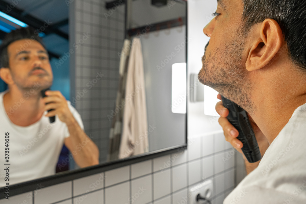handsome man is shaving his beard with trimmer machine in front of bathroom mirror - obrazy, fototapety, plakaty 