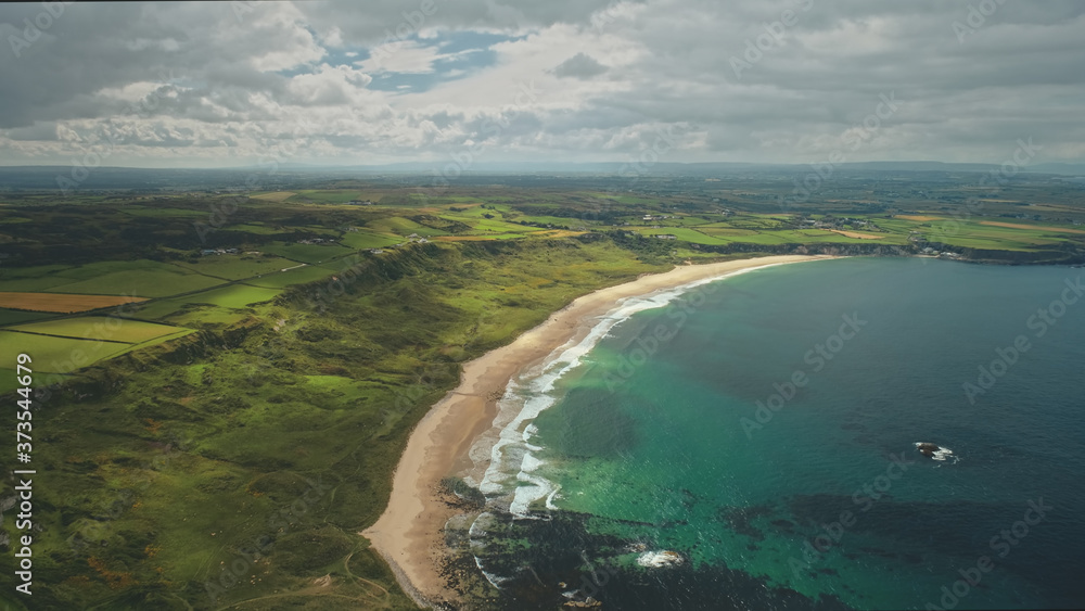 Beach aerial view panoramic landscape in Antrim County, Northern Ireland. Beautiful Atlantic Ocean coastline, green grass meadows and countryside stretch out to grey rain clouds.