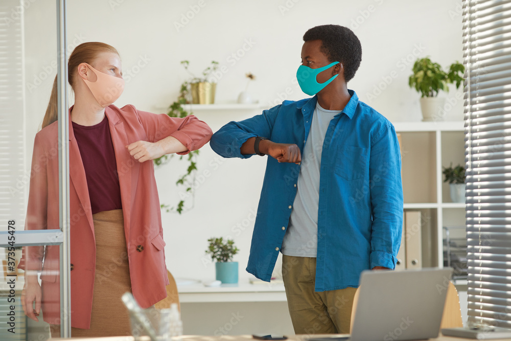 Obraz premium Portrait of young woman wearing mask bumping elbows with African-American colleague as contactless greeting while working in post pandemic office