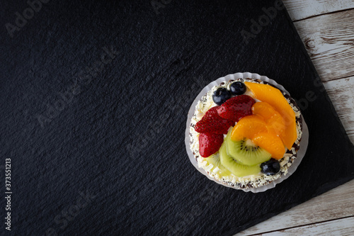 Fruit tart with strawberries, kiwi, blueberries and peach on black background placed on wooden table with space for copy text