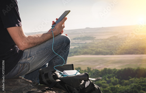 Man on a hike uses smartphone while charging from the power bank on the rock at dawn. Healthy lifestyle and communication.