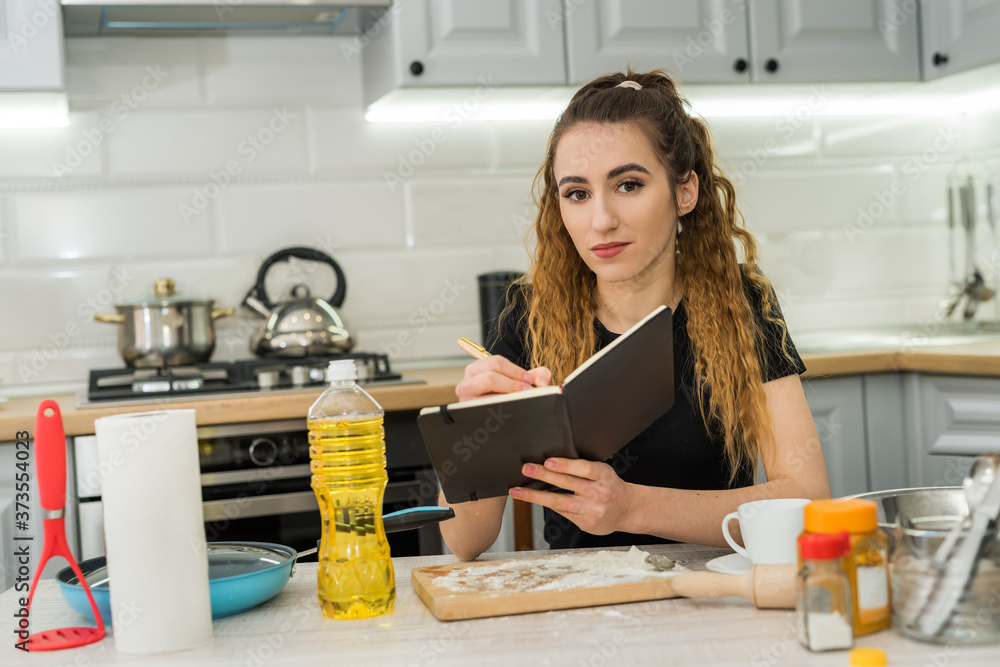 Young pretty lady reading notebook with recipe taste cake of flour, oil and other ingredient
