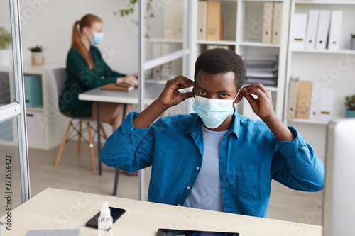 High angle portrait of young African-American man putting on face mask and looking at camera while working at desk in post pandemic office
