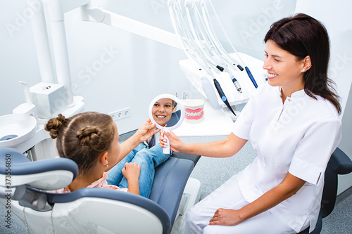 Little girl looking in mirror on healthy teeth after treatment, while sitting in a dental chair. Child's dentist communicate with little patient