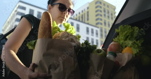 Woman Puts Paper Bags With Groceries For Week In Open Trunk Of Car. Buyer. Resident of Big City. Bought Food at Supermarket. View from Car Trunk. Woman in Sunglasses. Summer. City Street. Closes Trunk