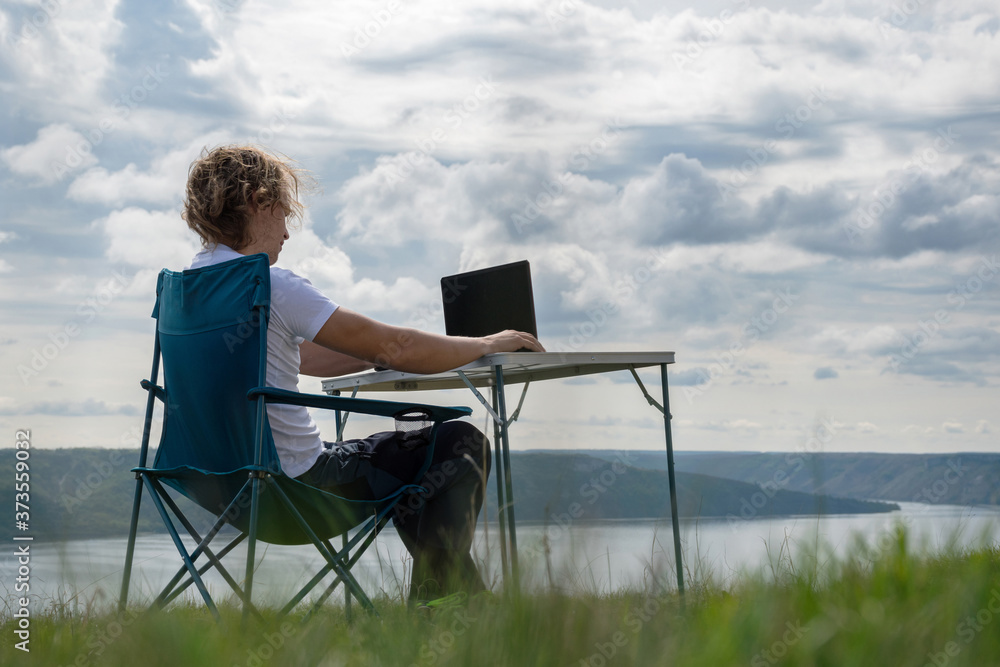 Remote work in nature. The guy sits on a chair at a table by the river and works on a laptop.