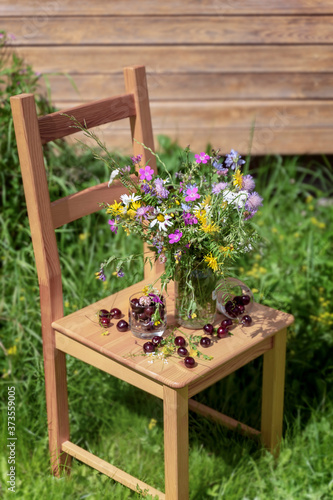 Bouquet of wildflowers and ripe cherries on a wooden chair in the grass, summer farm rustic concept. Natural background