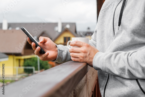 Man drinking coffee or tea on the balcony on the background of mountains and villas or pensions, Close up mans hand using his smartphone. Concept of holidays in countryside.