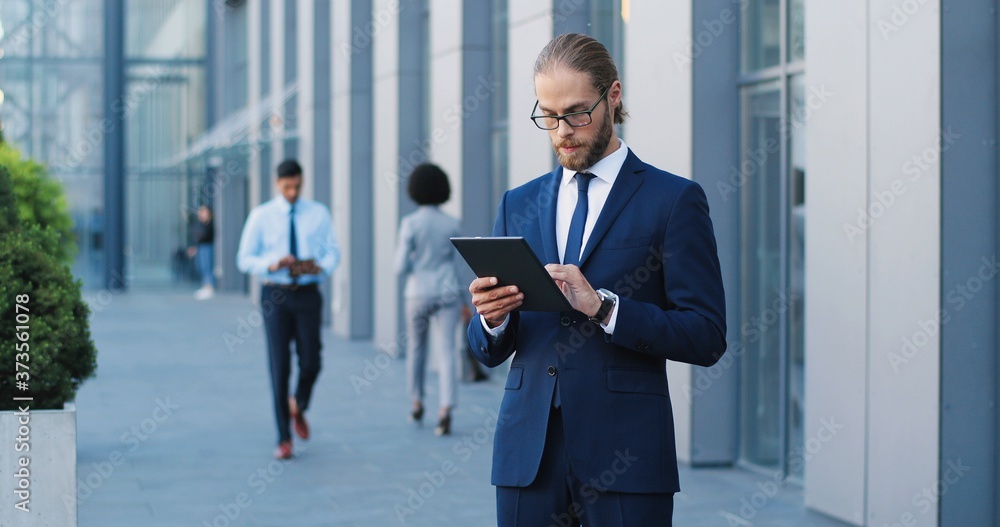 Handsome Caucasian young businessman in glasses, suit and tie using tablet device at street. Man in business style tapping and scrolling on gadget computer. People walking by. Pedestrians passing. Handsome Caucasian young businessman in glasses, suit and tie using tablet device at street. Man in business style tapping and scrolling on gadget computer. People walking by. Pedestrians passing.