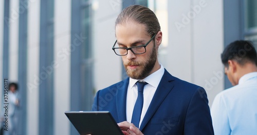 Handsome Caucasian young businessman in glasses, suit and tie using tablet device at street. Man in business style tapping and scrolling on gadget computer. Outdoor. Close up.