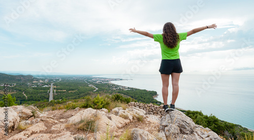 A girl at the top of a mountain, with her arms open looking at the sea