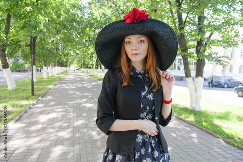 Red-haired girl in a big hat with red flower fields on a green alley in a summer park