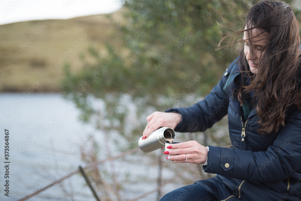 A woman sitting on a bench near a lake pours tea from a flask in a windy day in the Pentland hills regional park in Edinburgh, Scotland, United Kingdom A woman sitting on a bench near a lake pours tea from a flask in a windy day in the Pentland hills regional park in Edinburgh, Scotland, United Kingdom